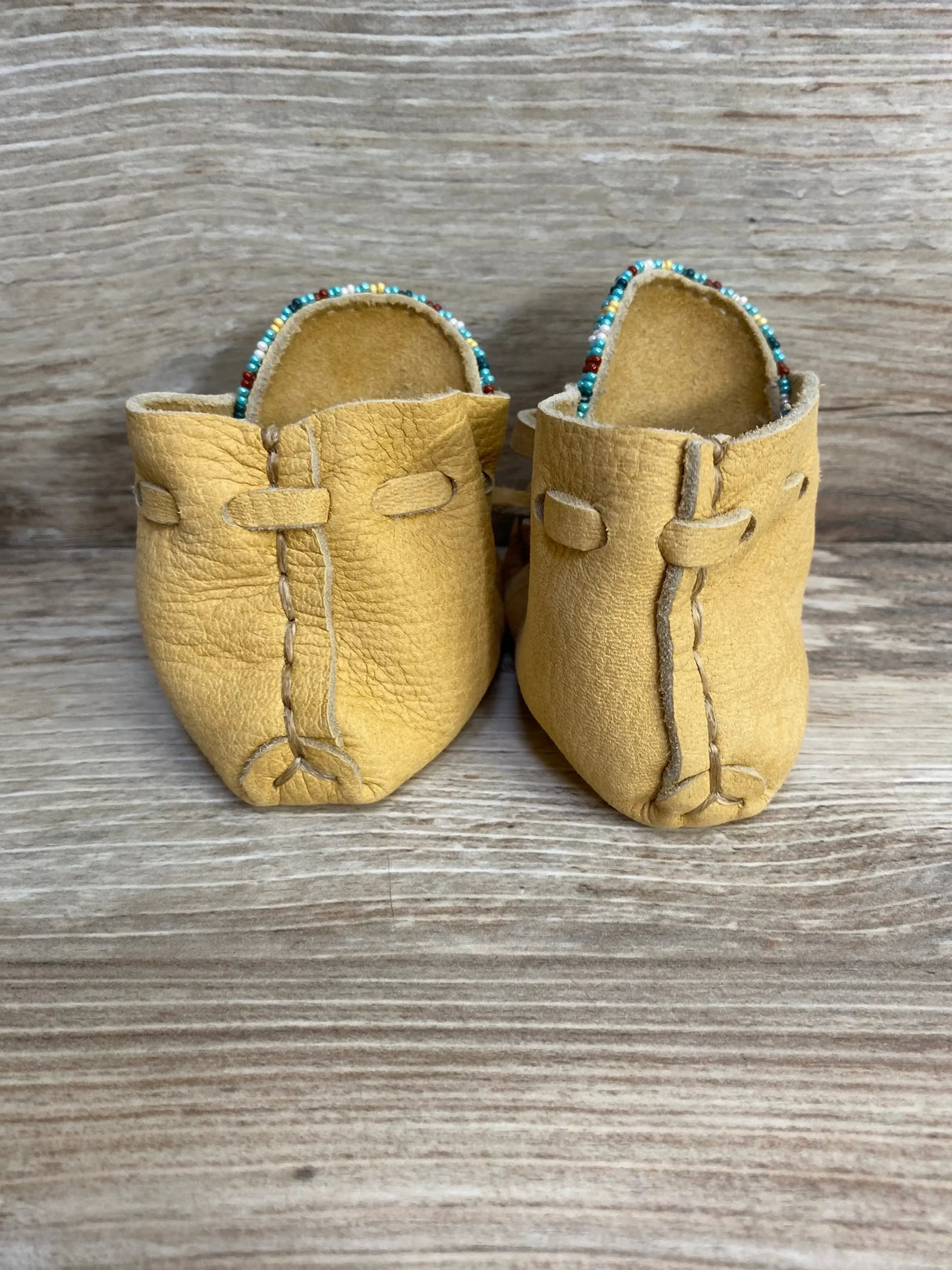 Front view of tan leather baby moccasin booties showcasing multicolor beaded trim and hand-stitched seams on wood background