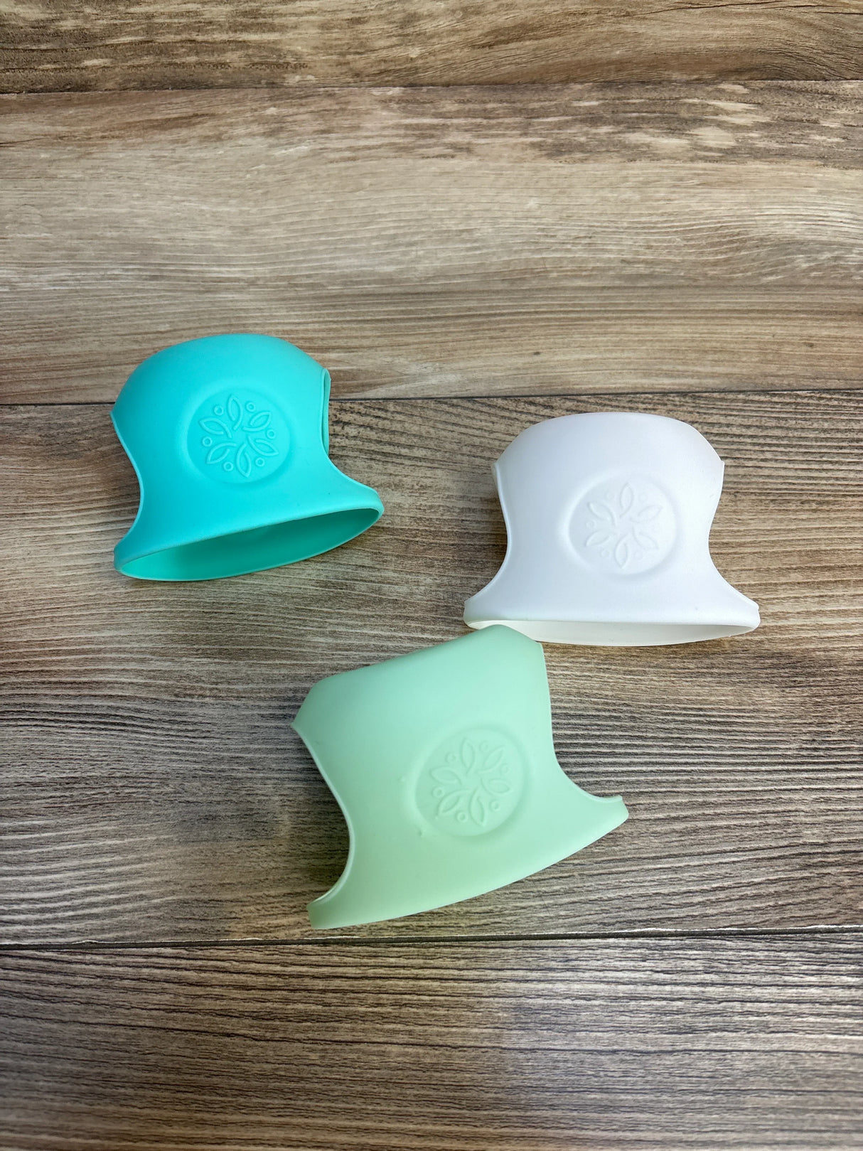 Three silicone baby bottle grips in teal, white, mint on wood — safe, non-slip infant feeding accessories