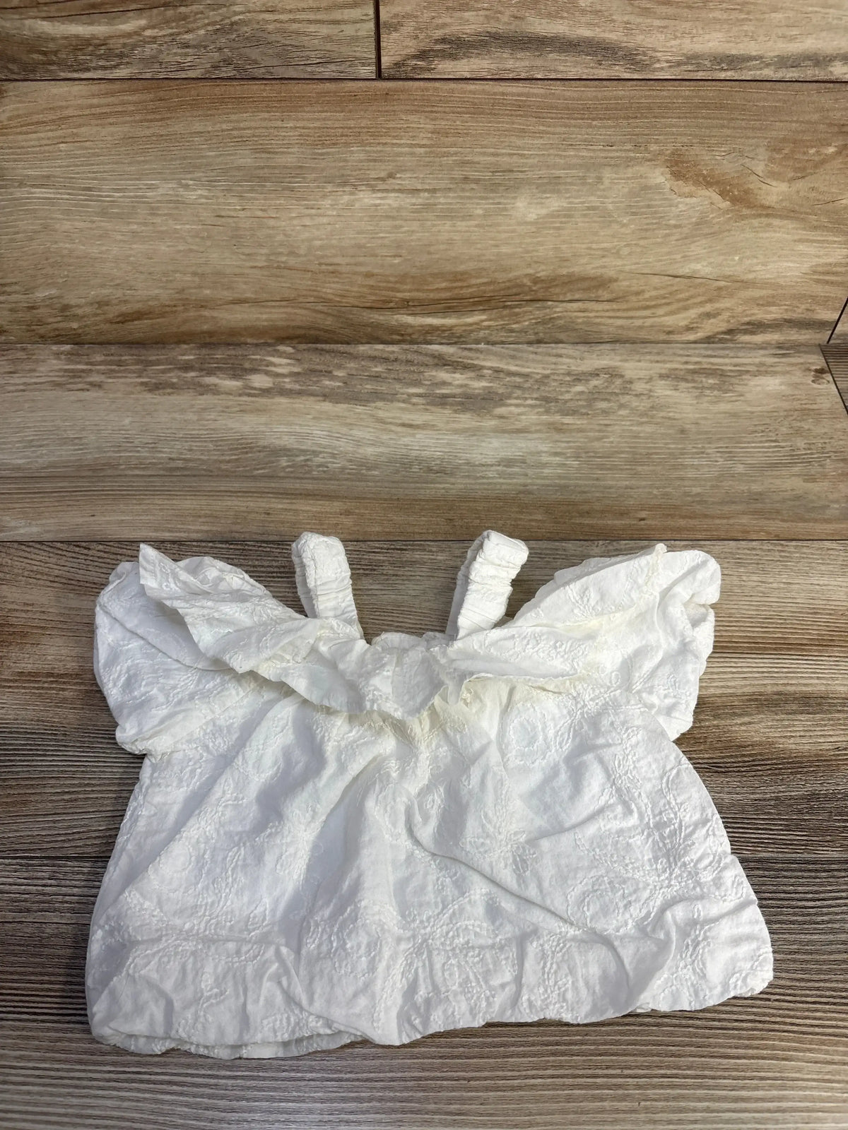 Flat lay of white Petit Main cold-shoulder top with floral embroidery, ruffle neckline, straps on wood background