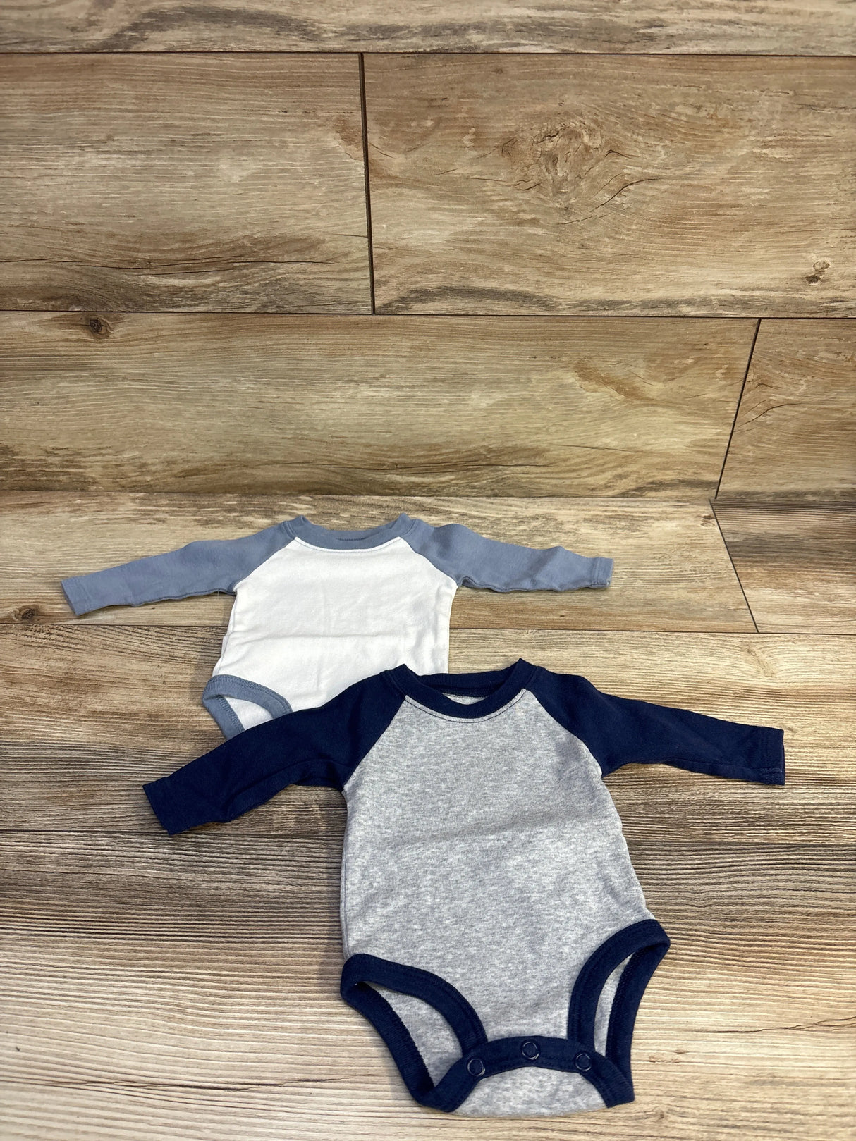 Top view of two Carter’s newborn raglan bodysuits in navy-gray and white-blue, showing three snaps on wood background