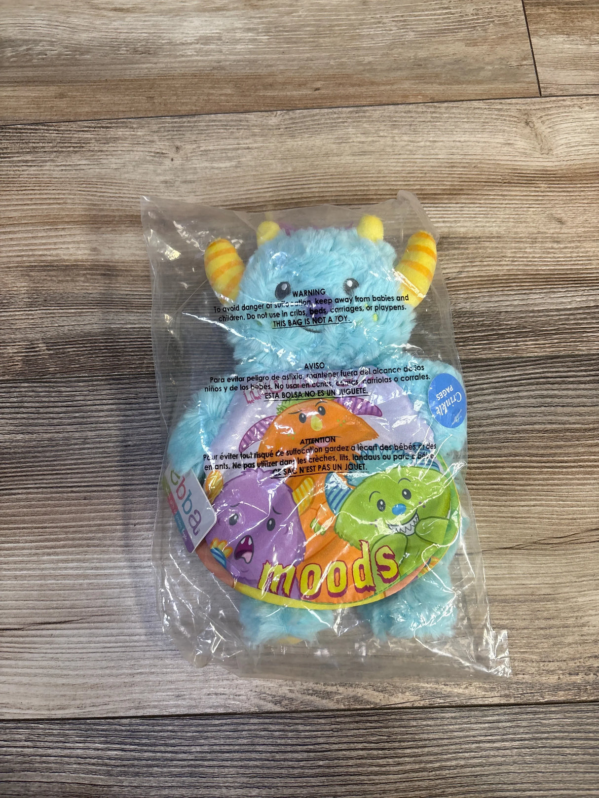 Top-down view of ebba Monster Moods plush book showing a smiling blue monster with yellow striped horns and ‘moods’ cover text on a wooden floor background