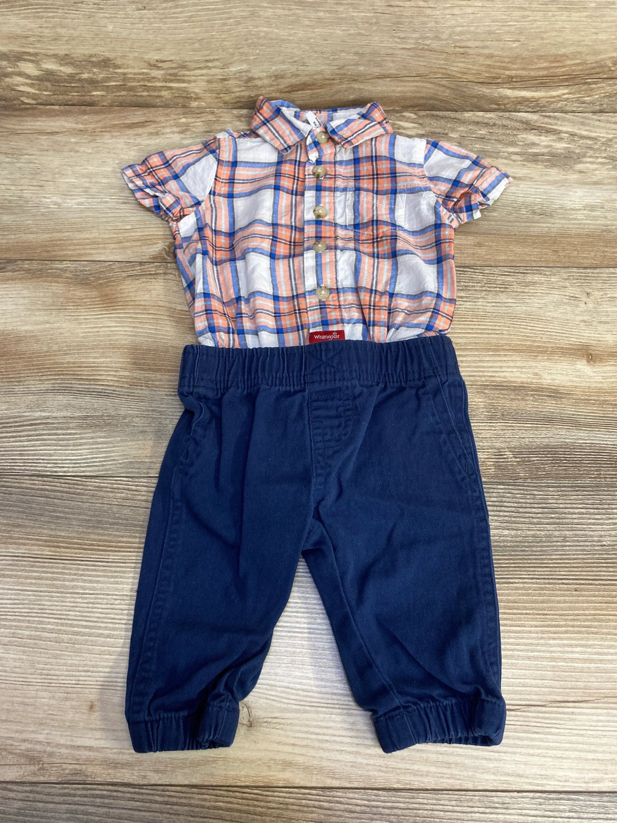 Flat lay of Wrangler two-piece baby outfit size 3M: orange and blue plaid short-sleeve button bodysuit with snap closure and navy elastic-waist pants on wood background