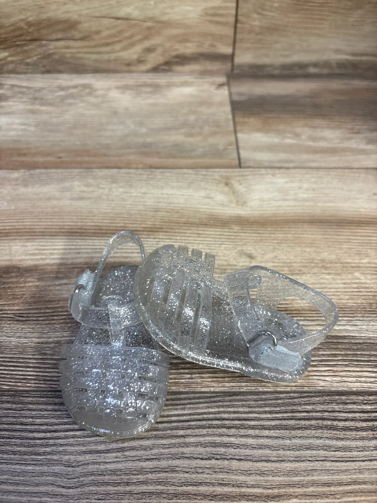 Angled top view of Old Navy toddler jelly sandals showcasing clear glitter PVC straps and ankle buckle with wood-grain background