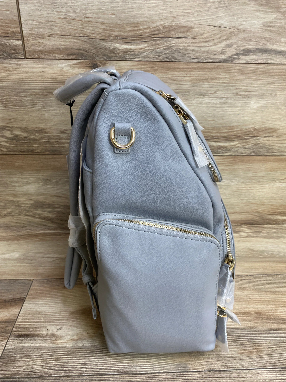 Light gray diaper bag backpack with side pocket and gold zippers on wood floor