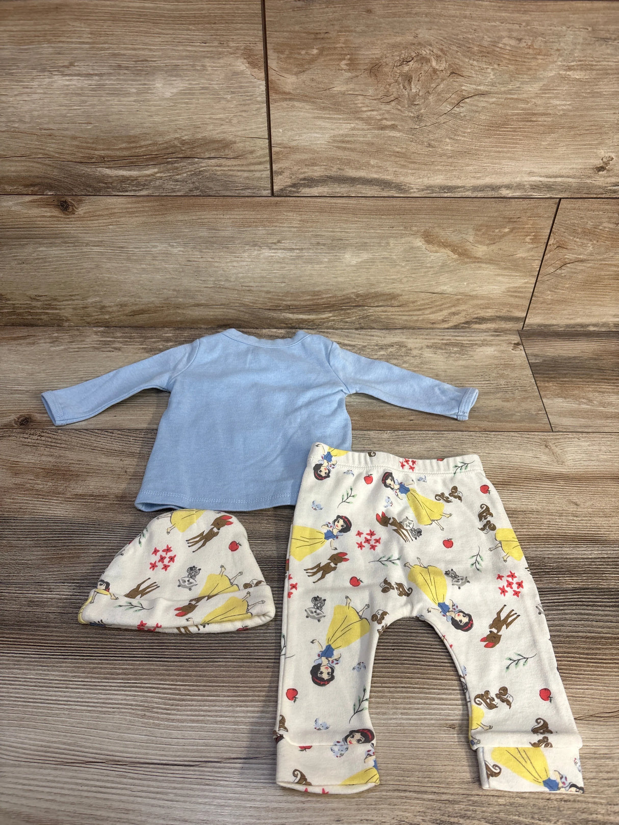 Top view of Disney Baby 3-piece set showing light blue shirt and cream Snow White print pants and hat on wood floor