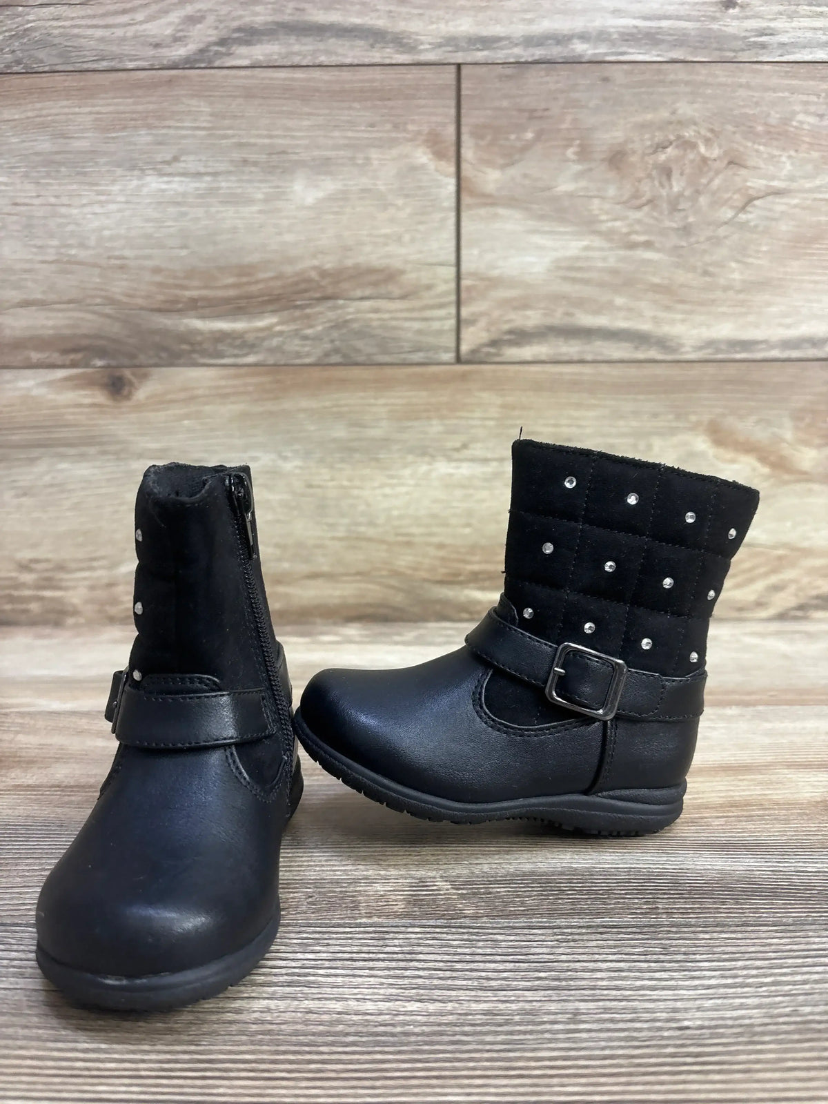 Pair of black toddler boots with side zipper, quilted upper adorned with rhinestones, buckle strap, on wooden floor