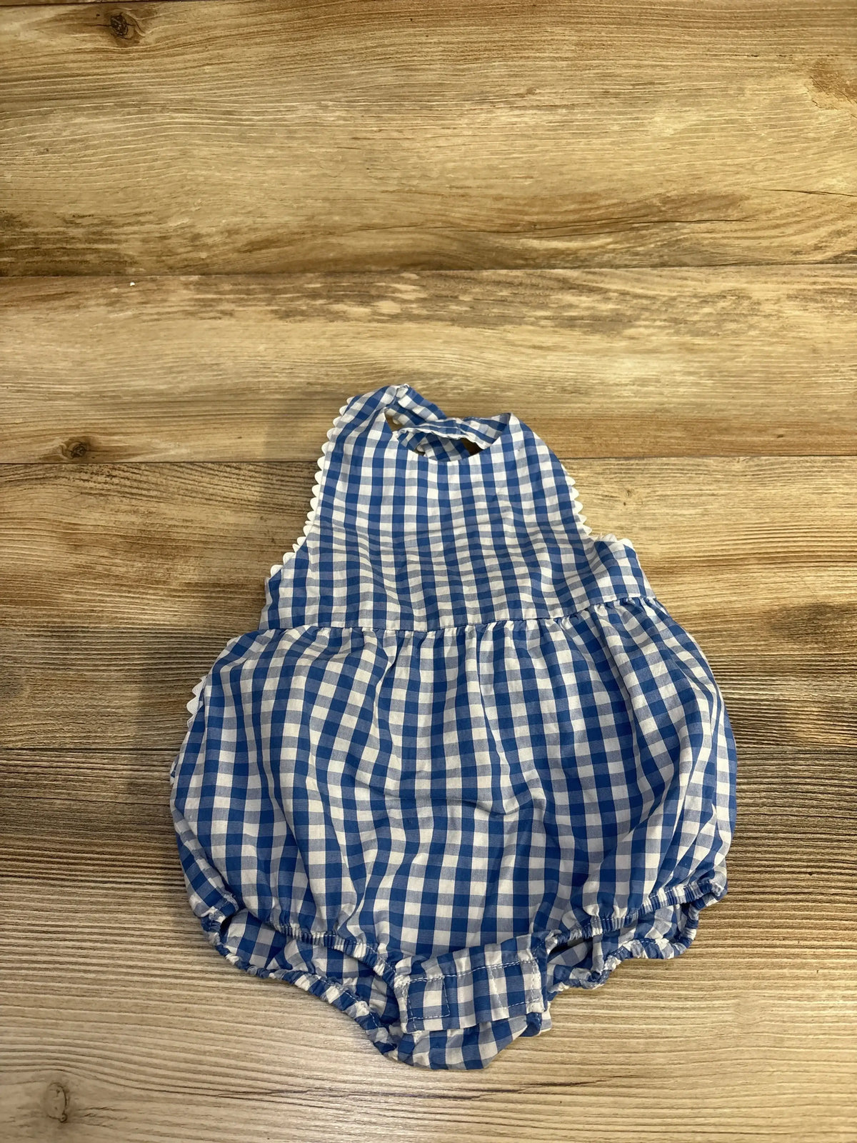 Flat lay of blue gingham bubble romper with ruffle scalloped trim, gathered waist, and elasticized ruffled leg openings on wood background