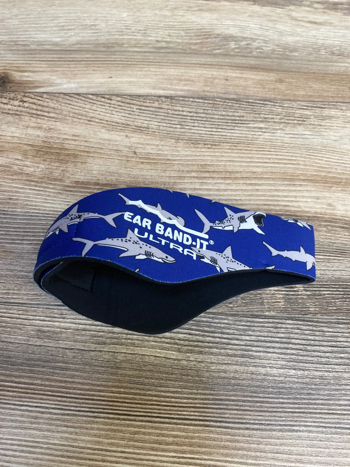 Side view of Ear Band-It Ultra swimming headband in blue with shark print showing padded black inner lining on wood surface