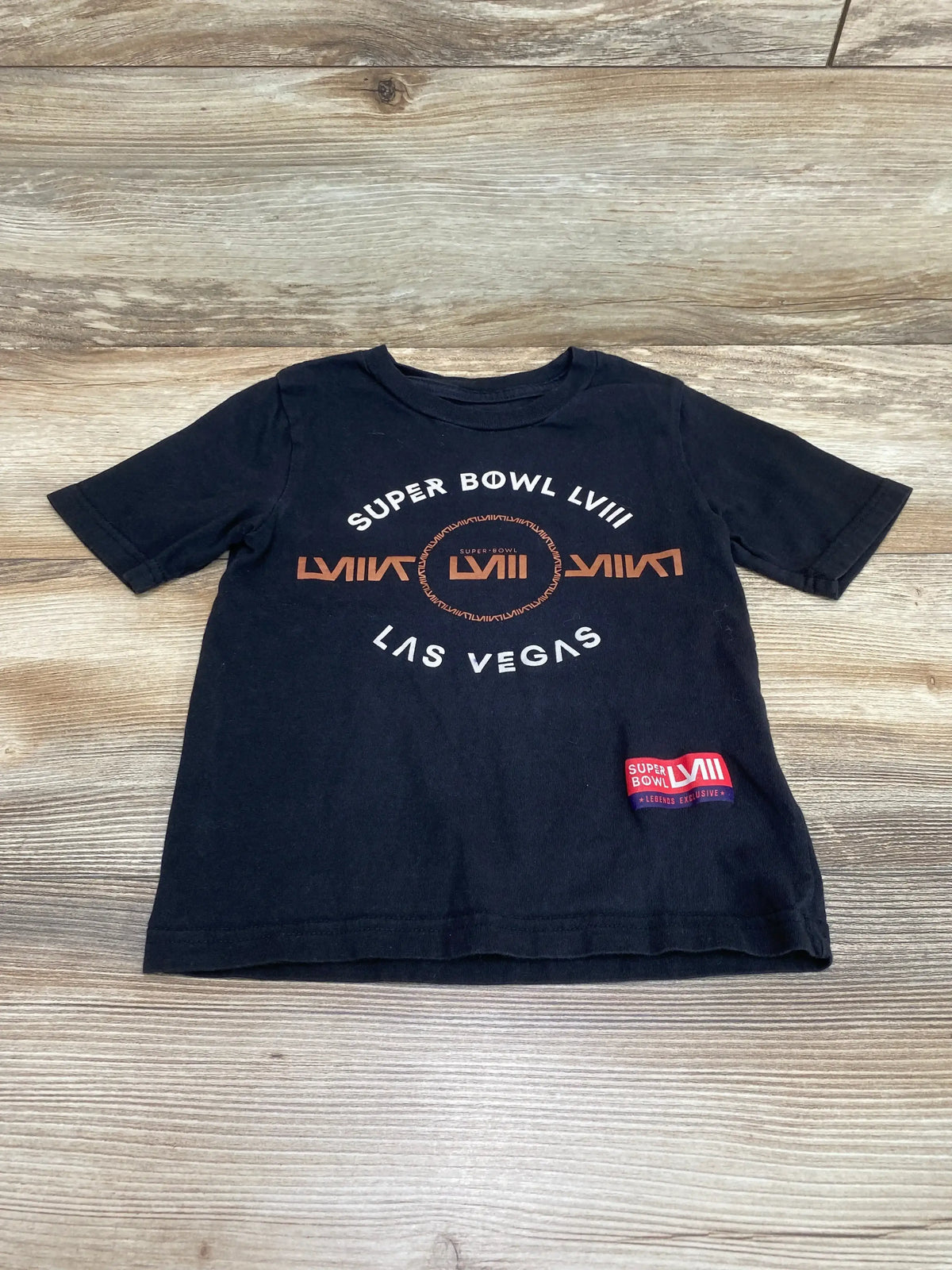 Flat lay black 4T T-shirt with 'Super Bowl LVII' and 'Las Vegas', central LVII logo and red patch on wood background