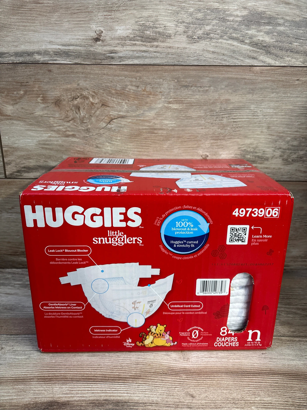 Front view of Huggies Little Snugglers newborn diaper box, 84 count, showing diaper illustration, wetness indicator labels and side window on wood background