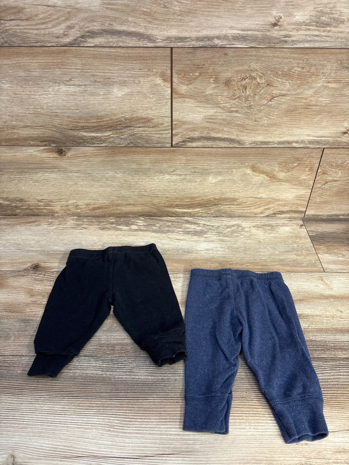 Two pairs of Simple Joys baby pants in black and navy blue on a wooden surface.