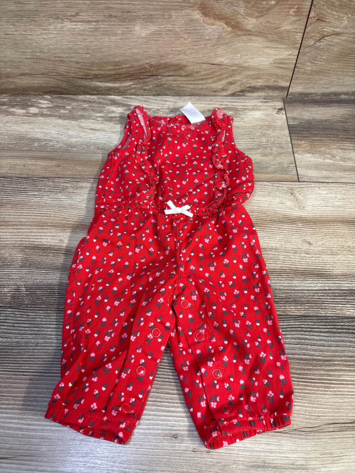 Front flat-lay of red floral baby jumpsuit (3M) showcasing ruffle shoulder straps, white waist bow, and snap leg closures on wood floor