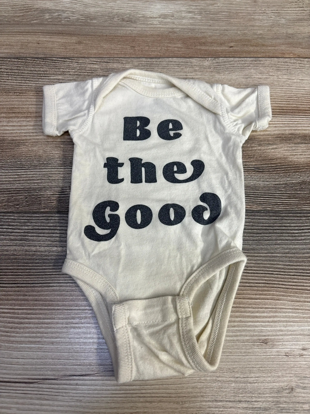 Cream baby onesie with black text 'Be the Good' on wooden surface