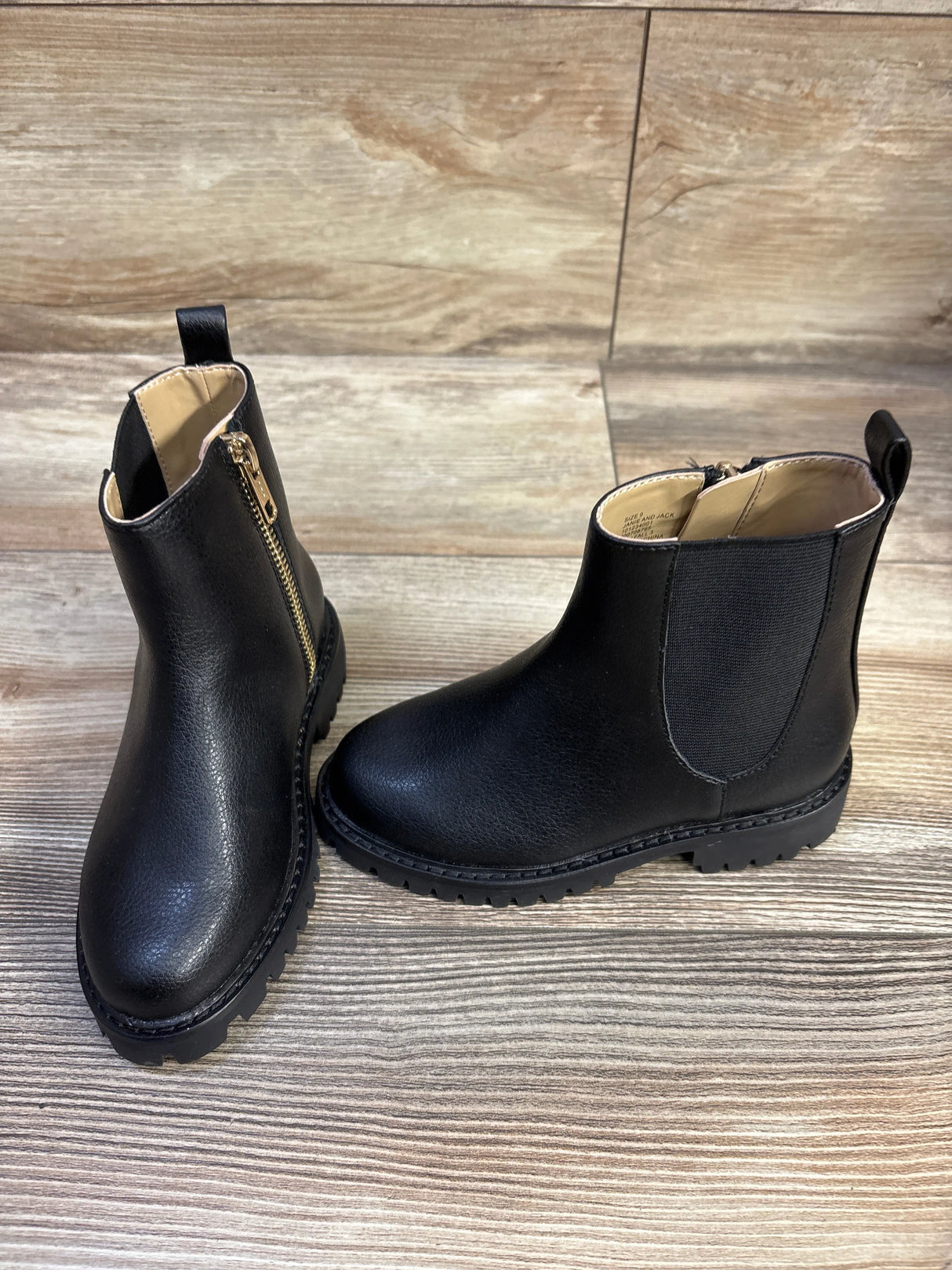 Front and side view of black children’s Chelsea boots showcasing gold side zipper, elastic side panel and lug sole on wood background