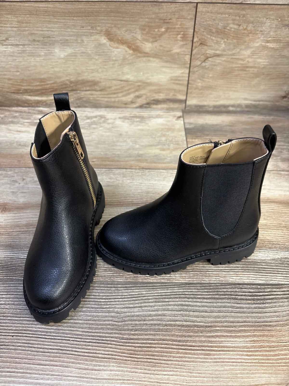 Front and side view of black children’s Chelsea boots showcasing gold side zipper, elastic side panel and lug sole on wood background