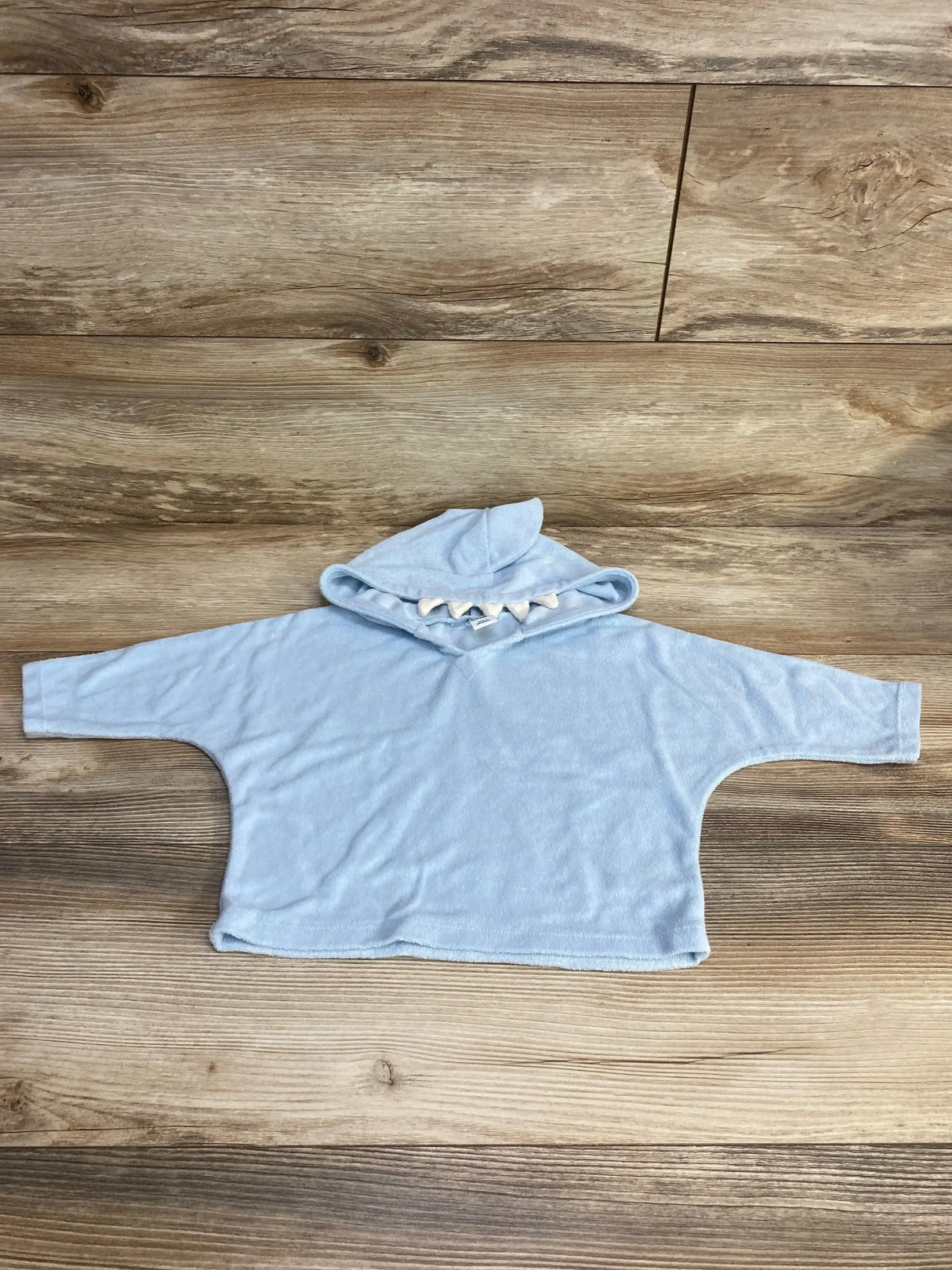 Flat lay of light blue terry baby swim cover-up hoodie (6–12 months) showing hood with shark-tooth trim on wood background