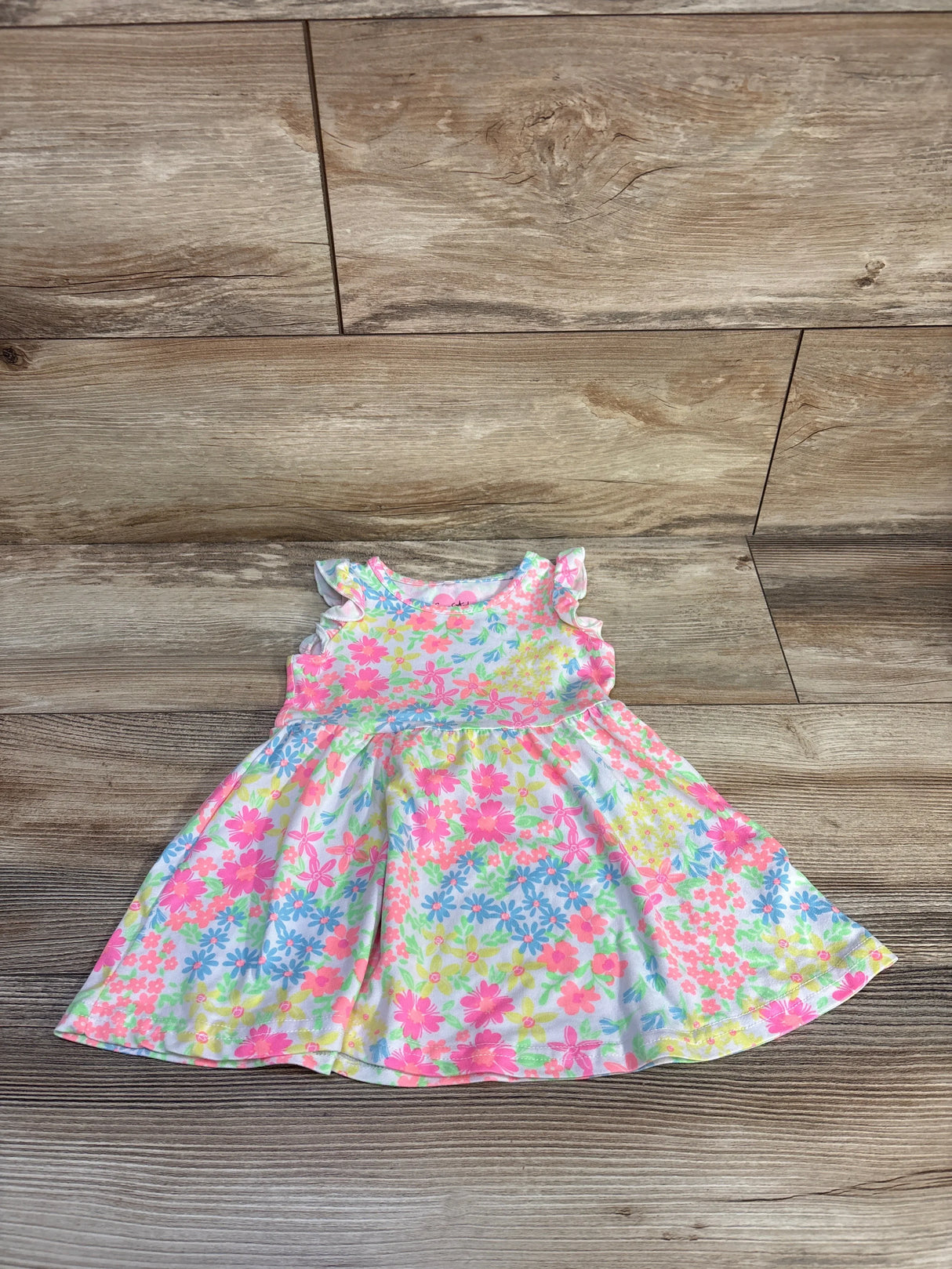Flat-lay front view of white floral toddler dress (12 months) showcasing ruffled cap sleeves and a gathered skirt on a wood floor background