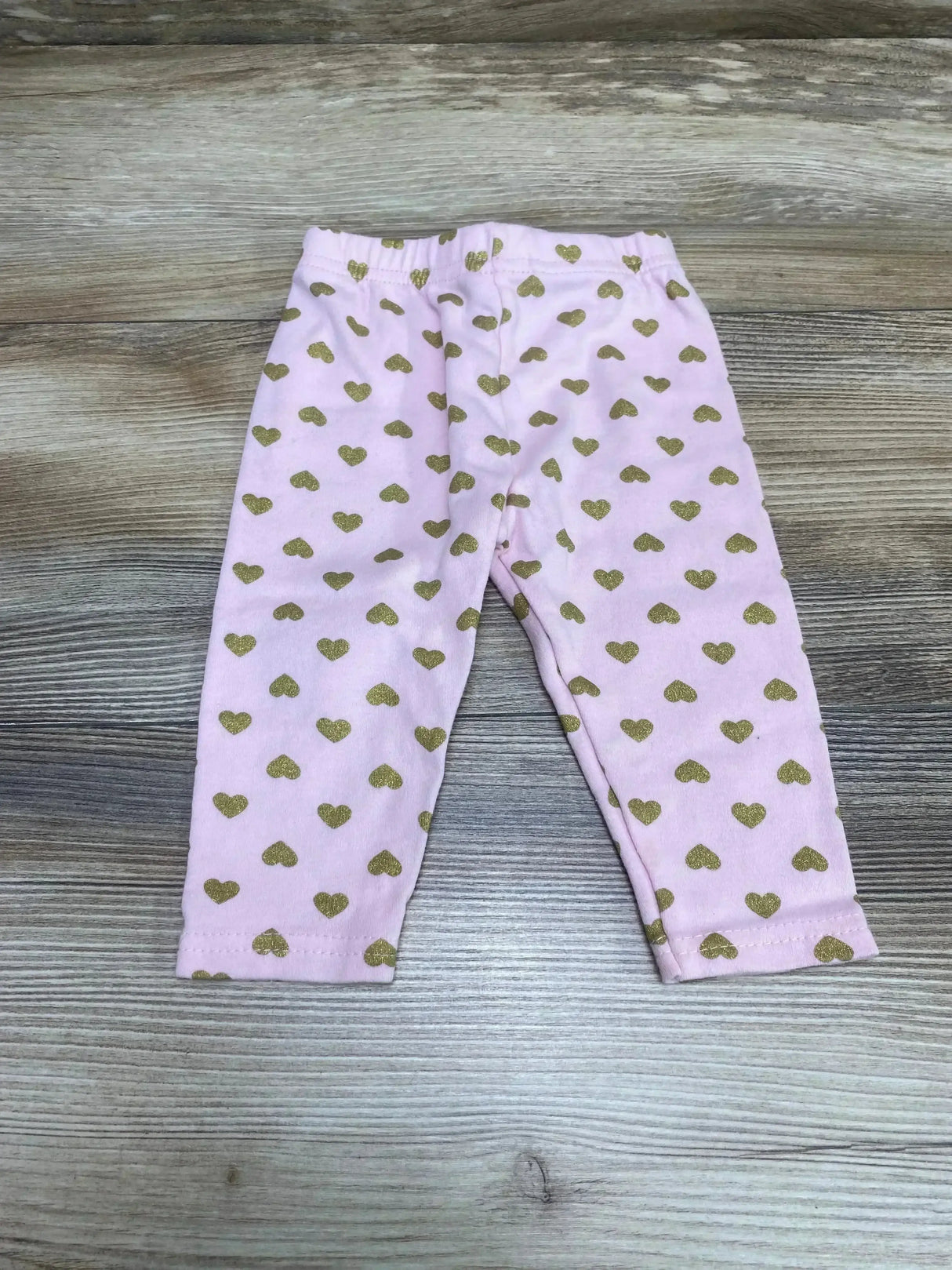 Flat lay of pink baby leggings (size 3–6 months) showcasing gold heart print and elastic waistband on light wood background