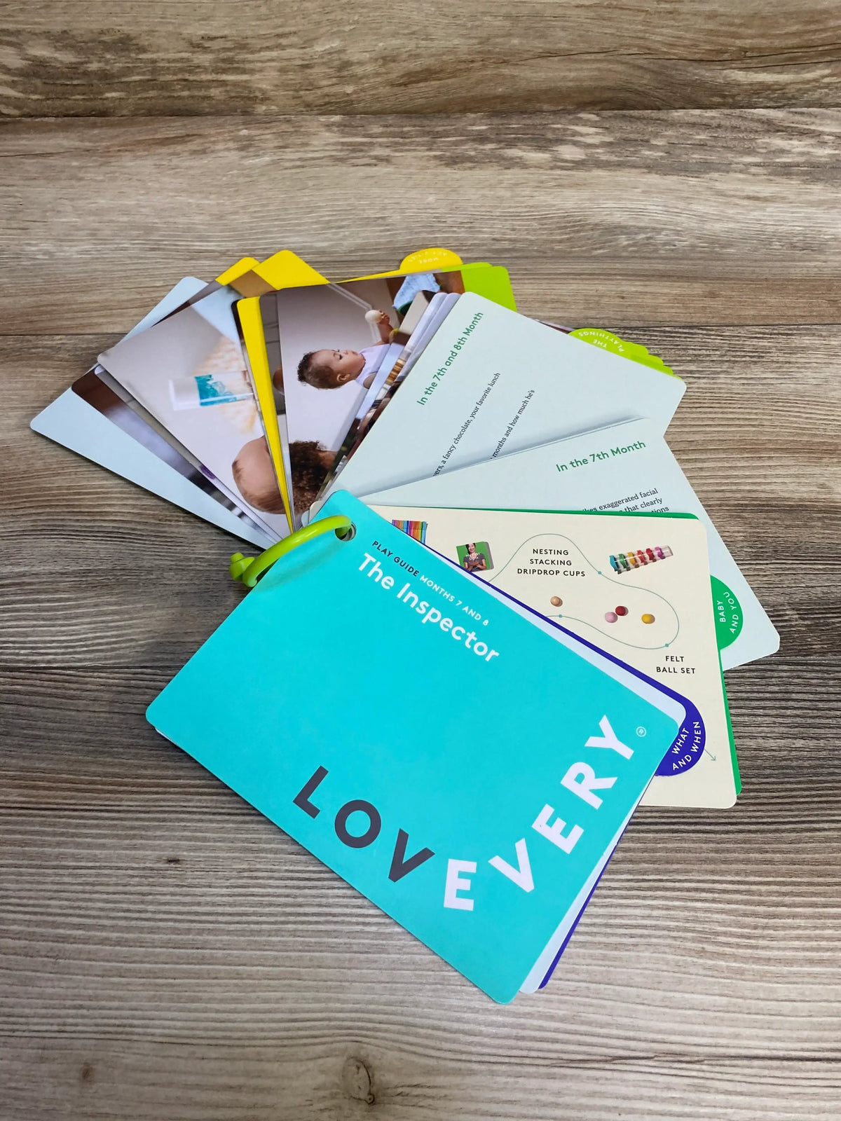 Angled view of Lovevery The Inspector play guide cards fanned on a wood tabletop, teal cover showing LOVEVERY logo and ring binding