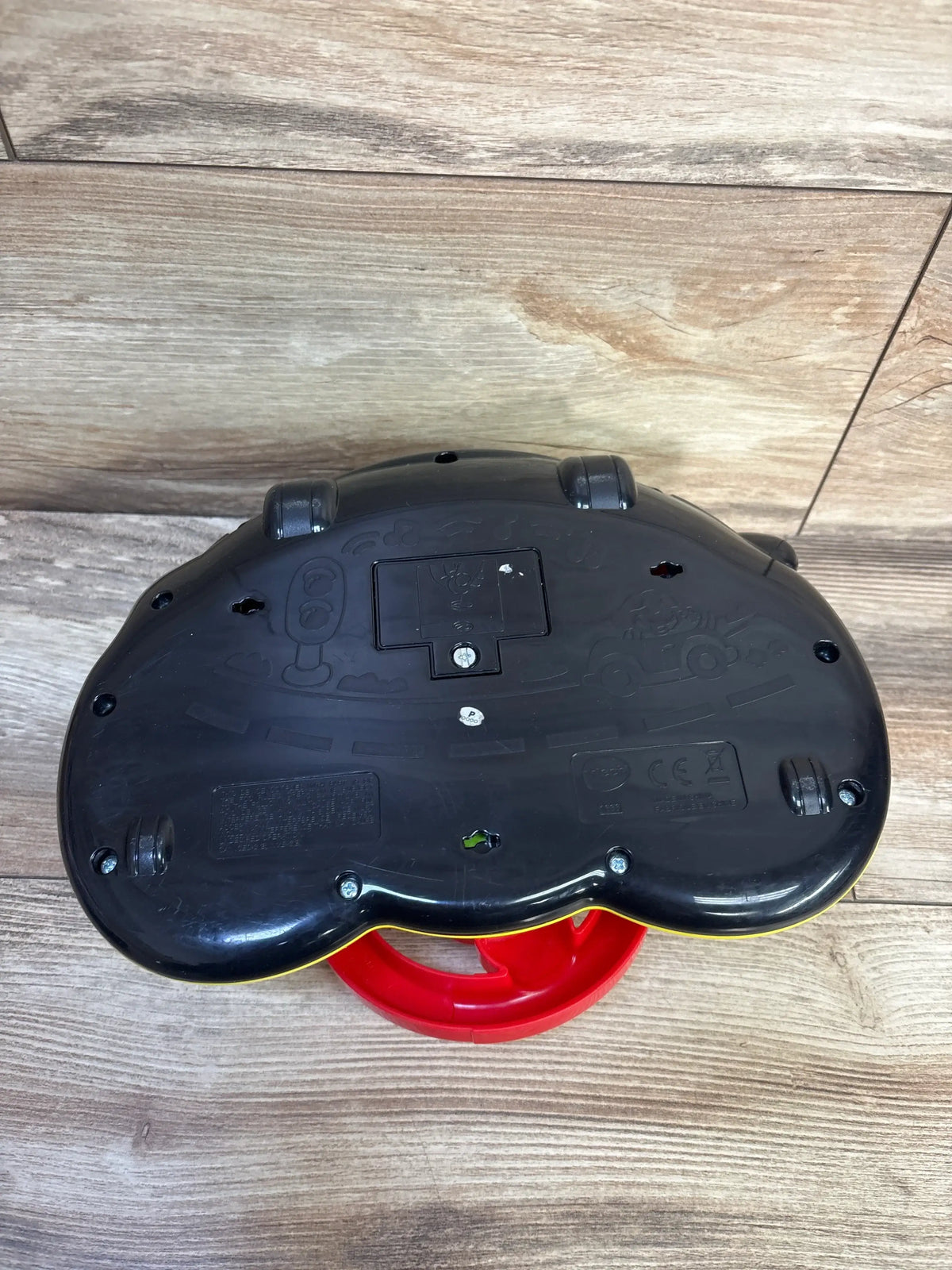 Bottom view of Vtech Turn & Learn Driver showcasing black underside, wheels, and battery compartment on wood flooring