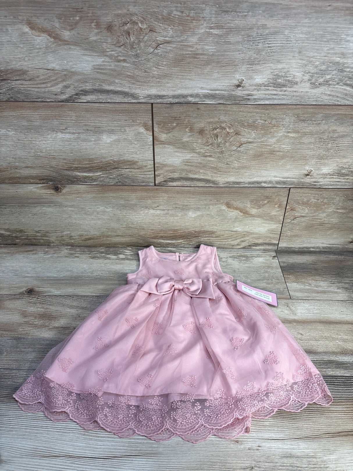 Flat lay of Katie M pink sleeveless tulle dress showcasing embroidered motifs, bow at waist, and scalloped lace hem on wood floor