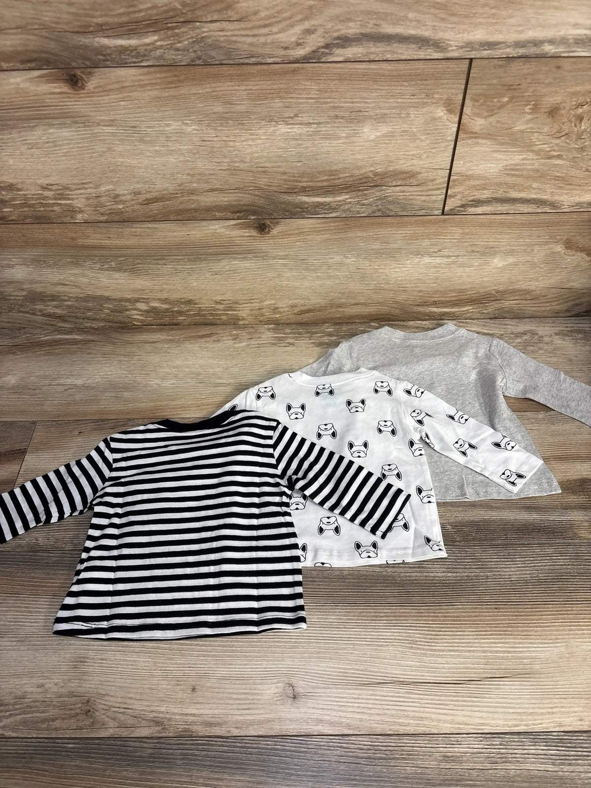 Front view of three long-sleeve baby shirts in grey, white dog print, and navy stripes on a wood floor