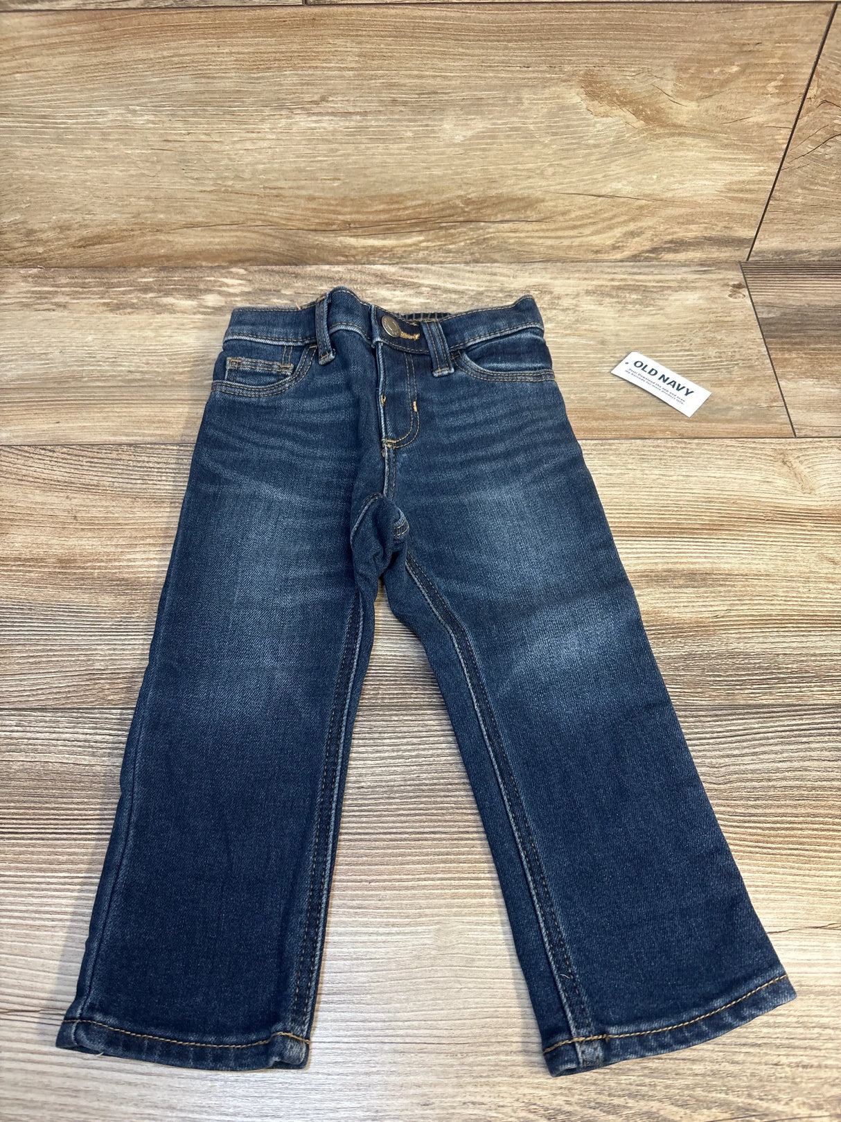 Flat lay of Old Navy toddler jeans, dark wash, size 18-24 months, showcasing straight legs and faded whiskering on wood floor background