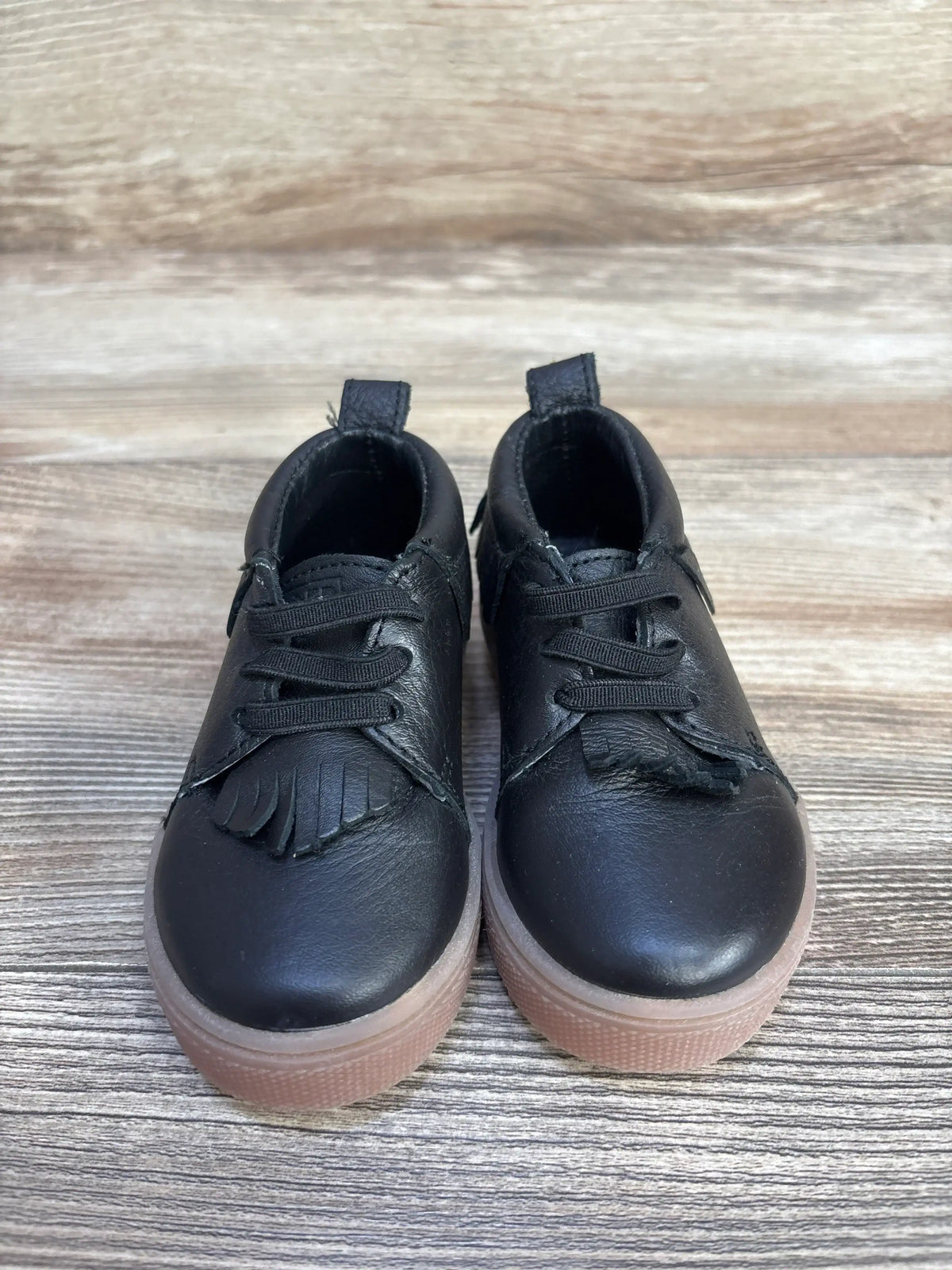 Front view of black Freshly Picked slip-on mid toddler sneakers size 5C showing fringe tongue and gum rubber soles on wood