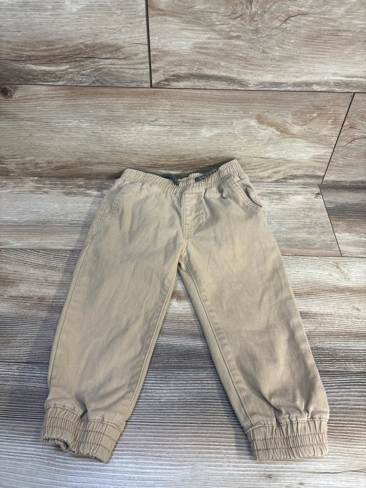 Beige kids jogger pants with elastic waist and cuffs, laid on wood floor