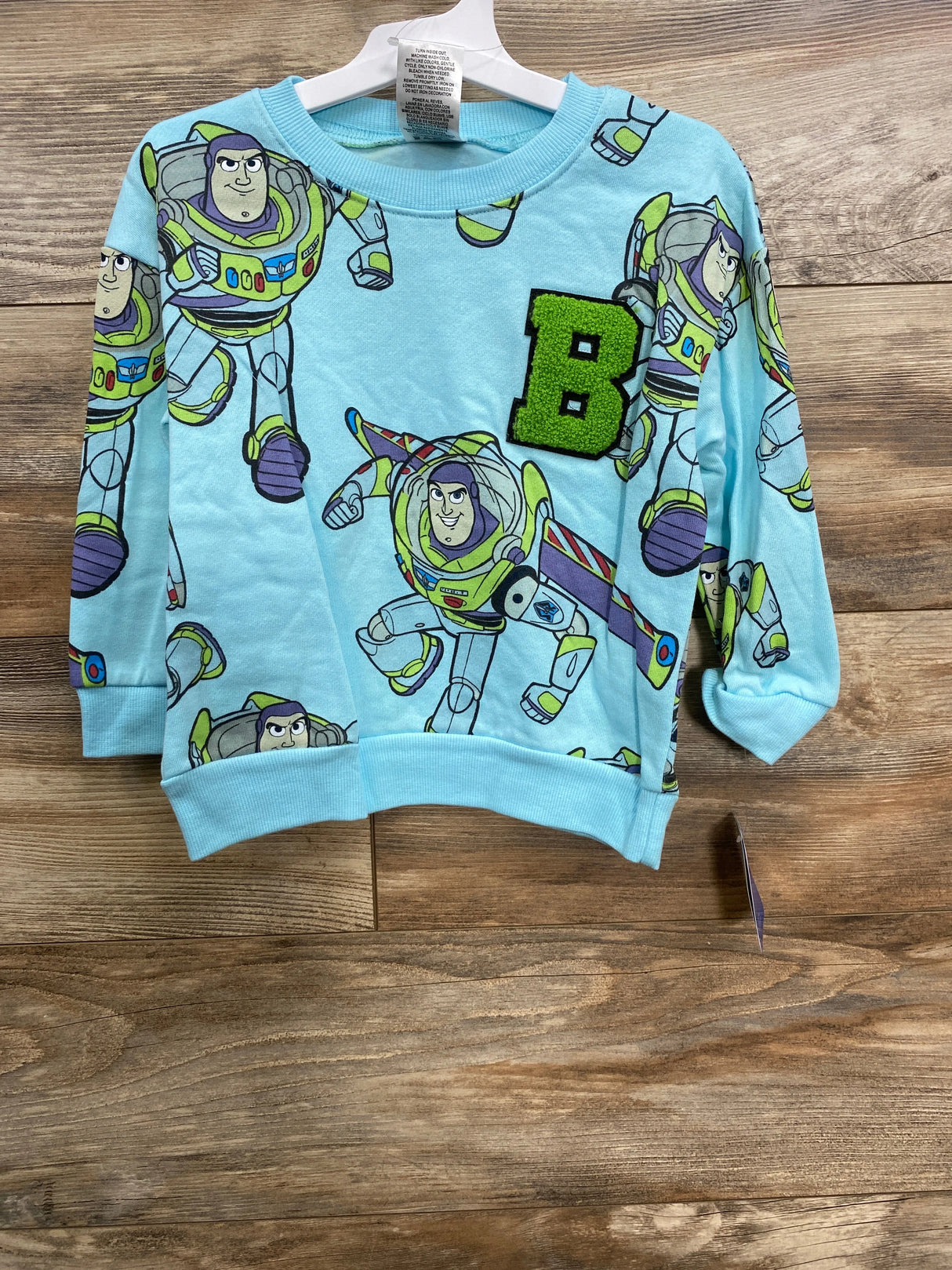 Light blue kids sweatshirt featuring Buzz Lightyear print and green textured letter B on wooden floor