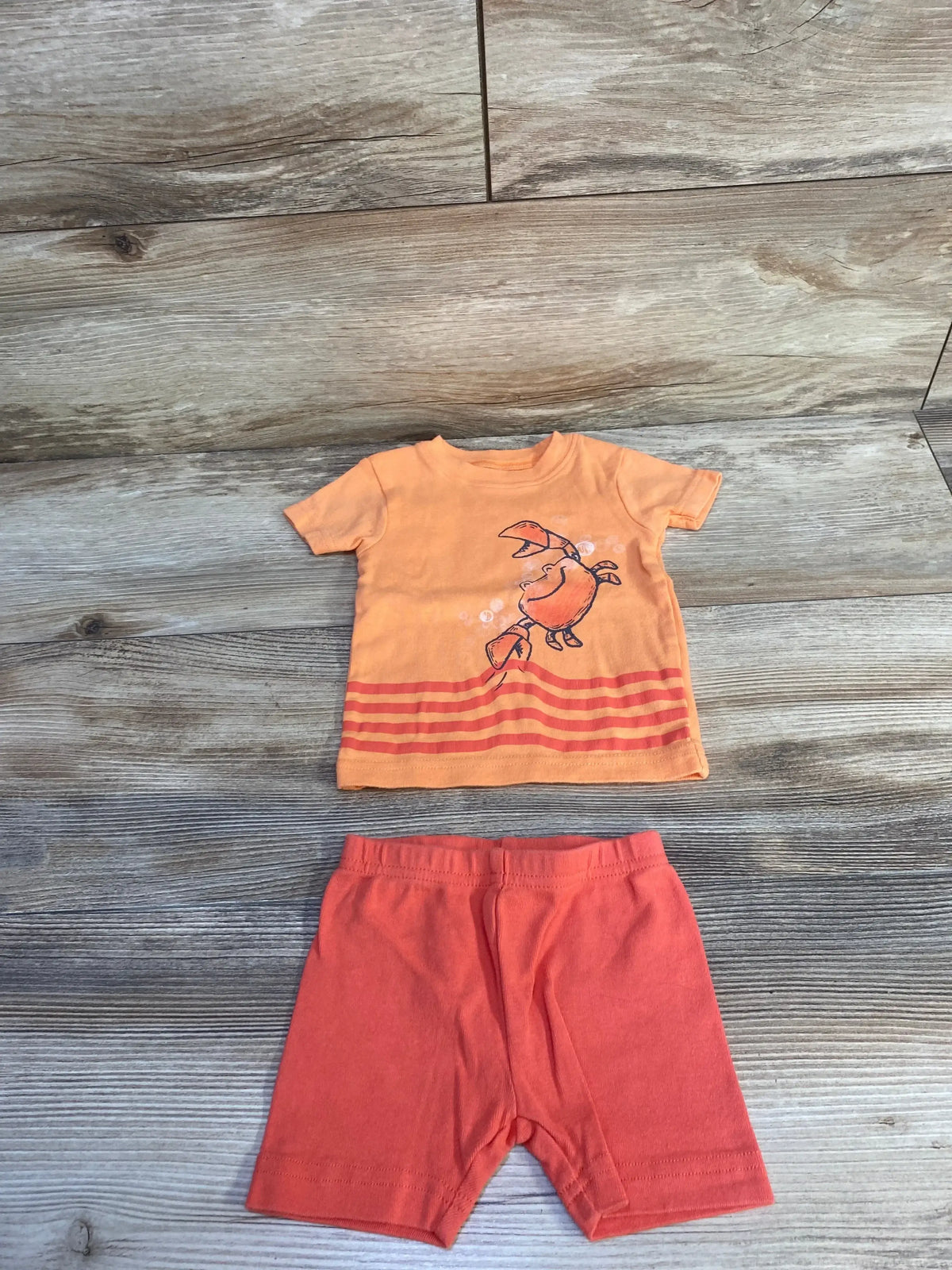 Flat-lay front view of orange 2-piece pajama set with crab graphic and striped hem, matching coral shorts on wood background
