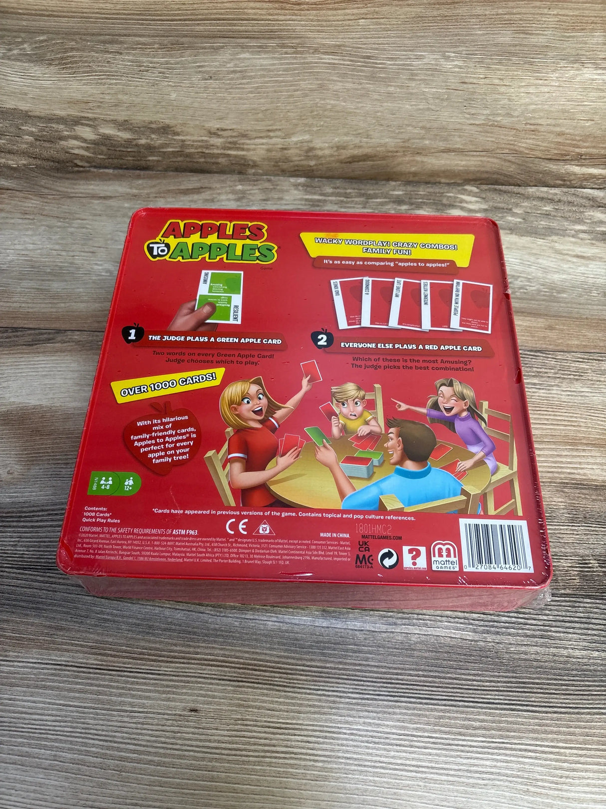 Top-down view of Apples to Apples game box back showing red artwork, gameplay instructions and an Over 1000 Cards badge on a wooden table