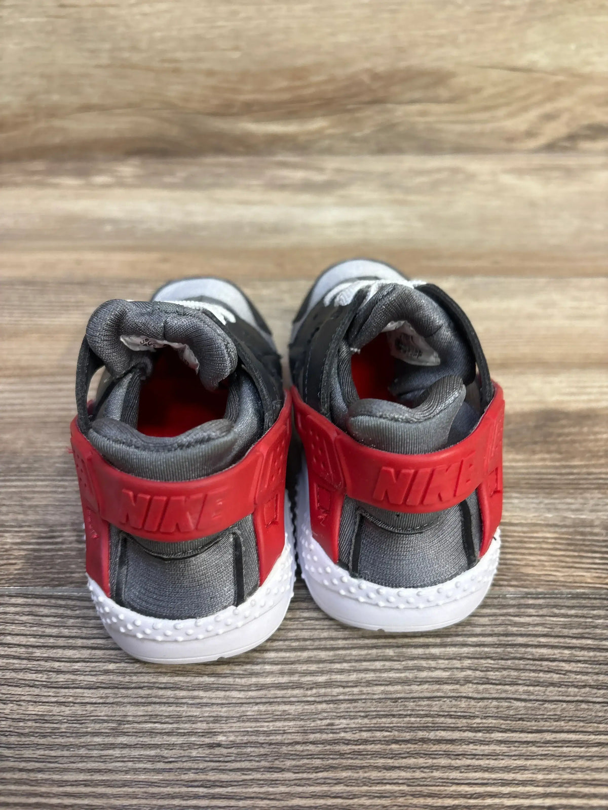 Rear view of Nike Huarache toddler sneakers with red heel strap embossed NIKE, gray upper and white sole on wood surface
