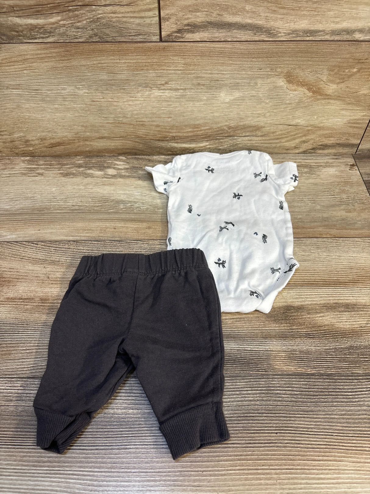 Flat lay of newborn 2-piece set: white animal-print bodysuit and black elastic-waist pants on wood floor
