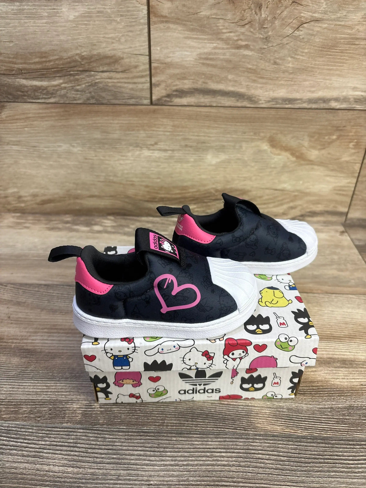 Side view of Adidas x Hello Kitty Superstar slip-on toddler sneaker, black with pink heart and white shell toe on Adidas box