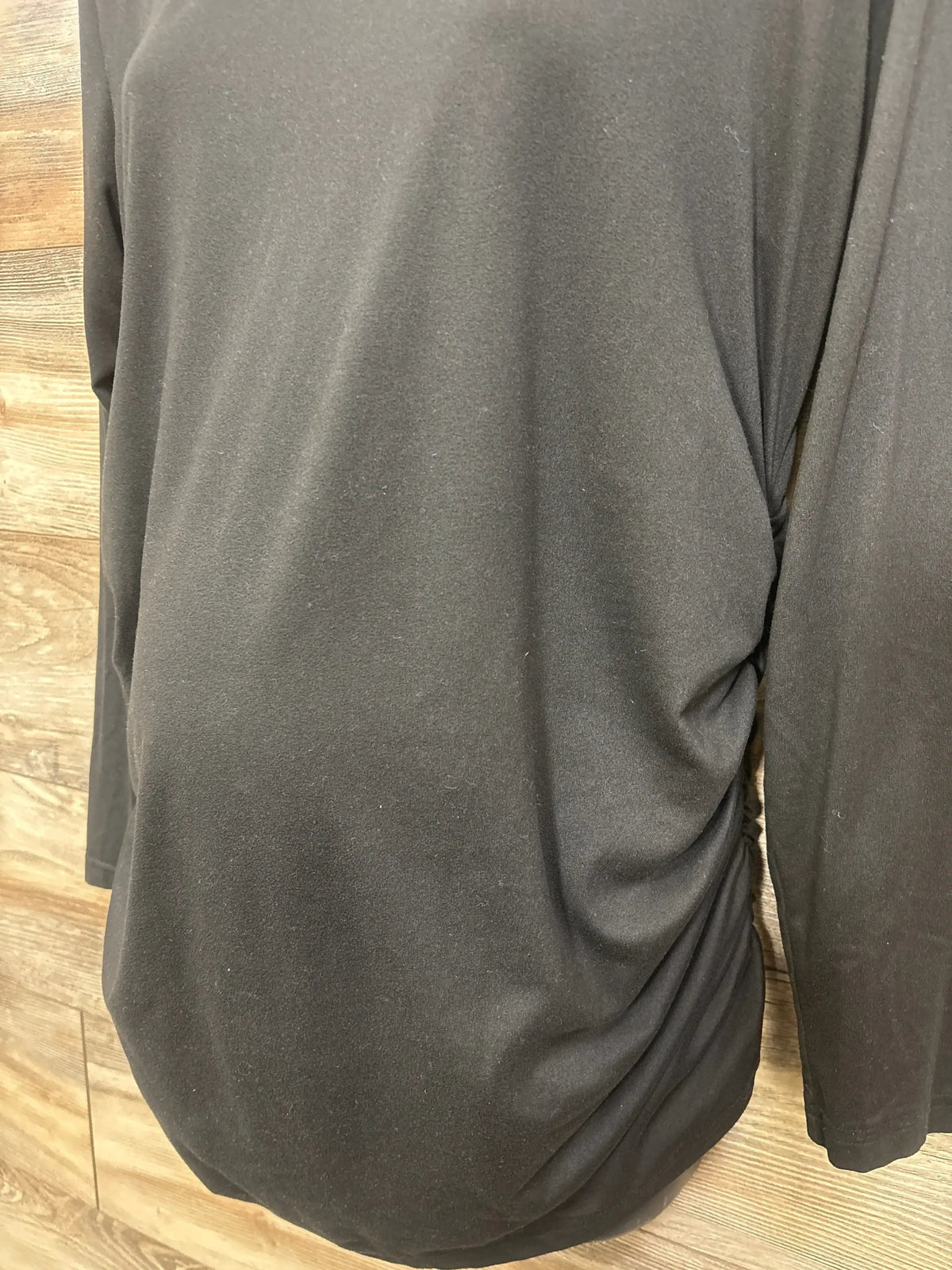Close-up back view of Ekouaer black ruched long-sleeve maternity top showing side ruching on wood-panel background