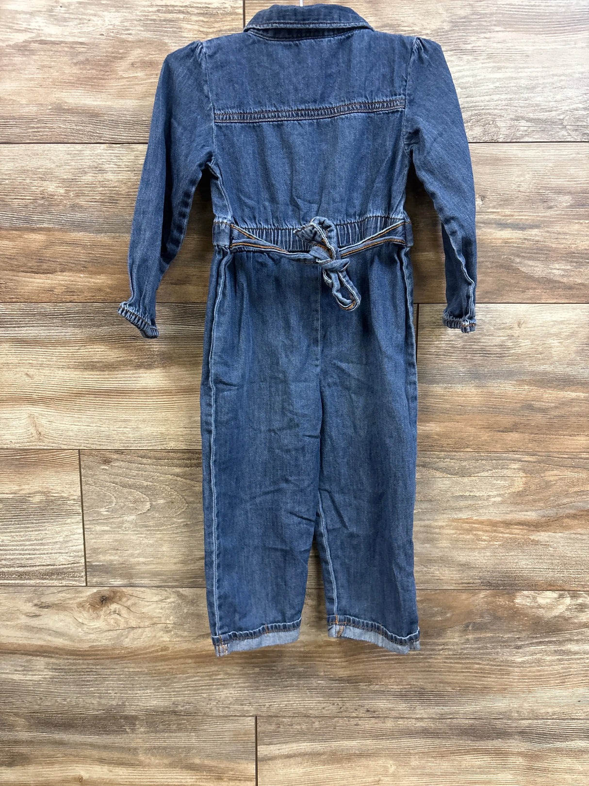 Back view of Oshkosh chambray polo coverall size 3T showing elastic waist with tied belt and long sleeves on wood background