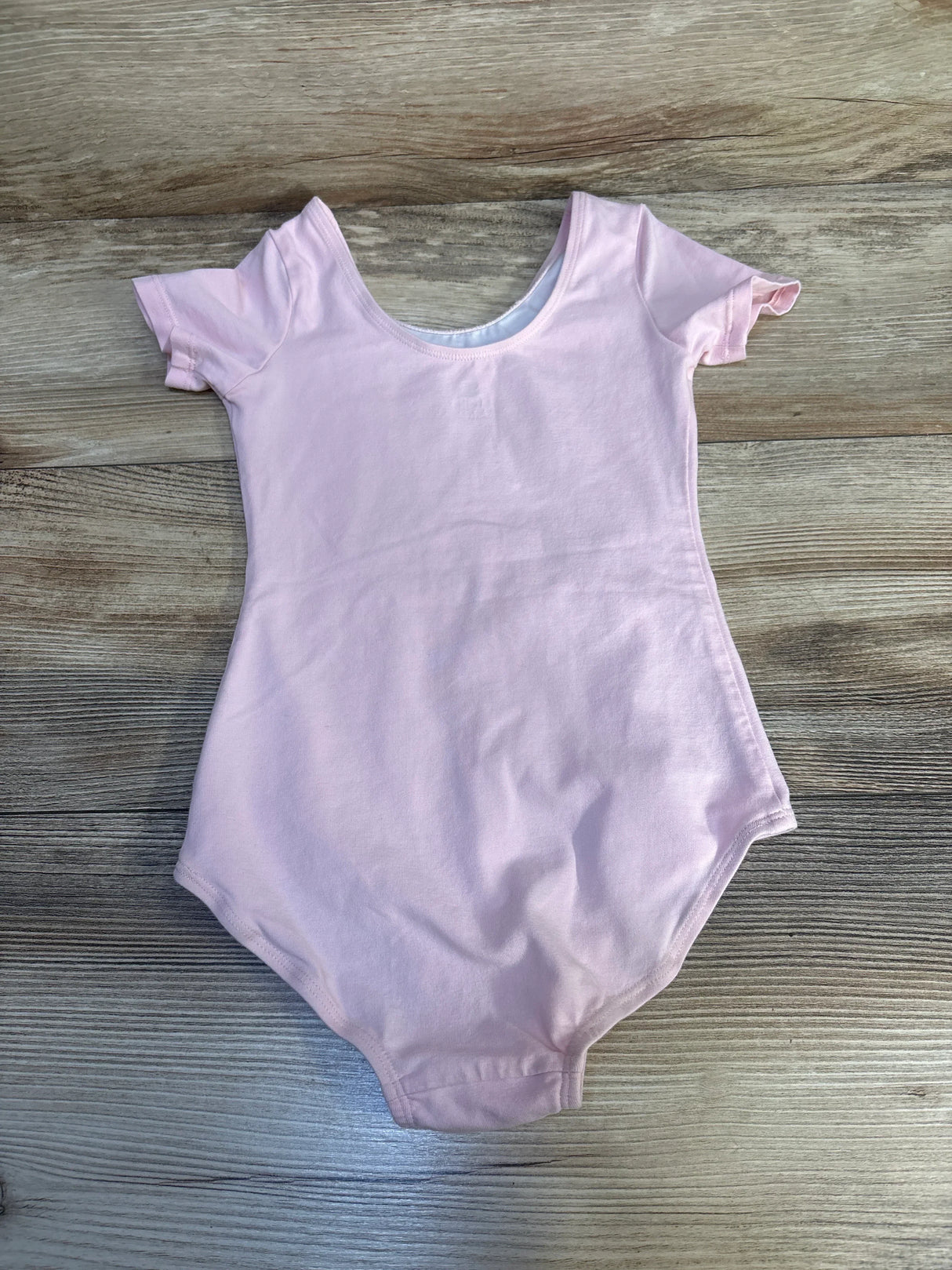 Flat lay of pink short-sleeve gymnastics leotard size 3T showing scoop neck and high-cut leg openings on wood background