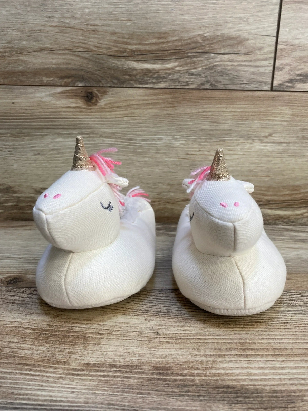 Pair of white unicorn baby slippers with pink accents and gold horns on wooden floor
