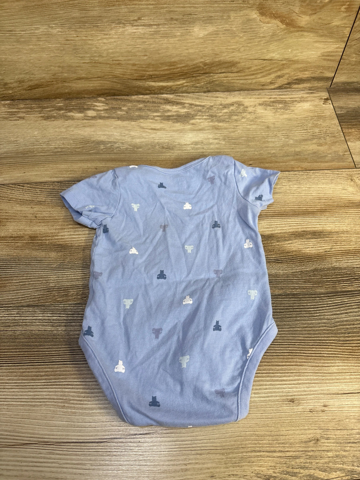 Flat lay of Baby Gap blue short-sleeve bodysuit showcasing small bear-head print on light blue cotton with wood background