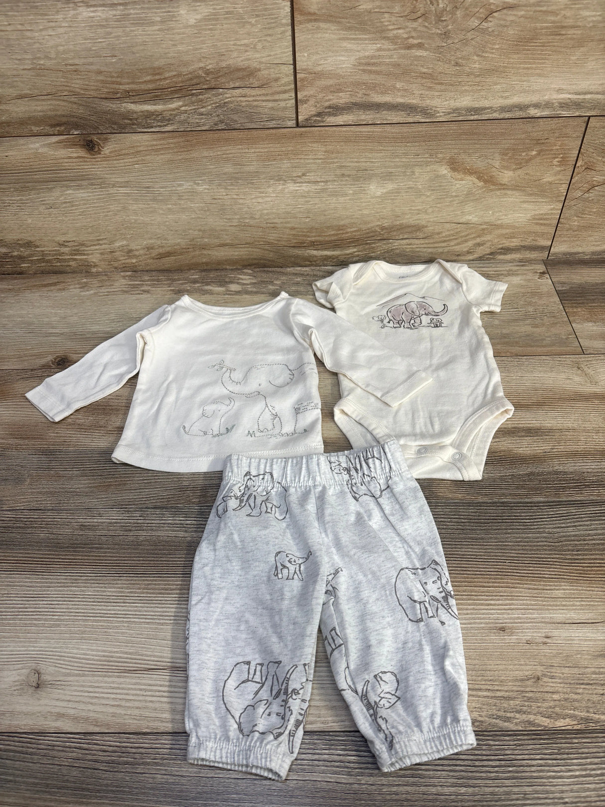 Flat lay of Carter's 3-piece ivory baby bodysuit set with elephant prints on top, bodysuit, and gray pants on wood floor