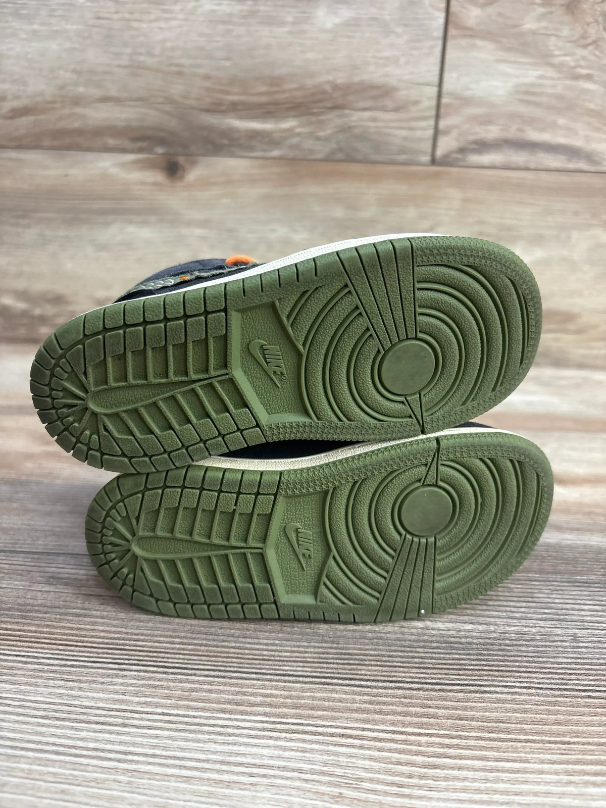 Bottom view of Air Jordan 1 Mid SE Craft TD sneakers showcasing olive rubber outsoles and tread on a wood floor