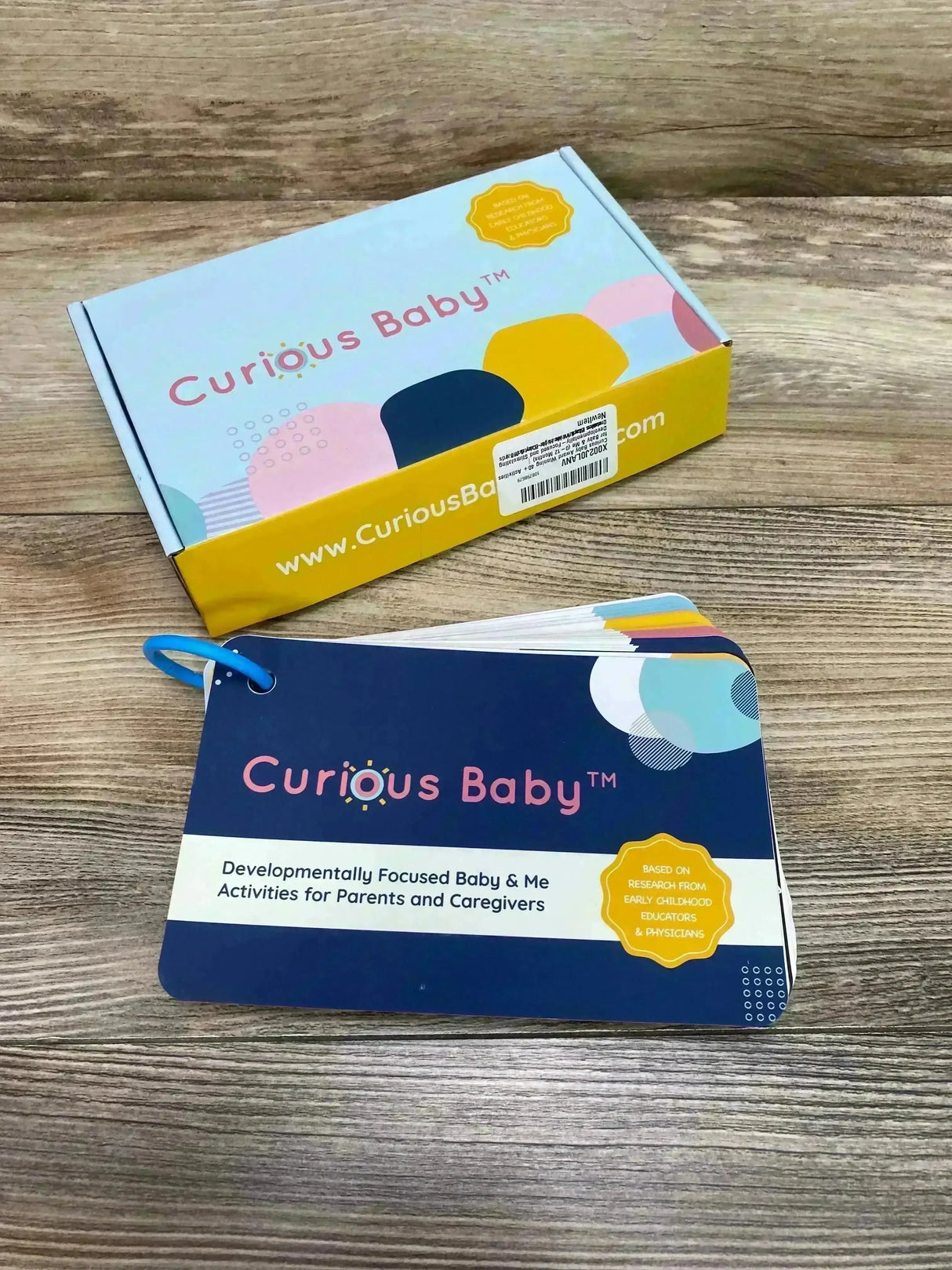 Angled overhead view of Curious Baby activity cards, a ring-bound navy deck labeled Curious Baby, with matching box on wood