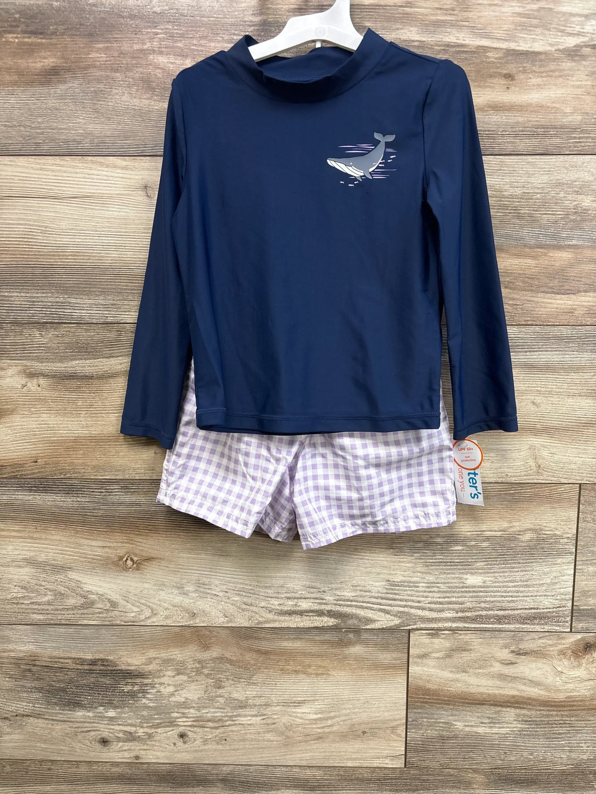 Front view of navy 2pc whale rash guard set: longsleeve top with whale graphic and lilac gingham shorts on wood background