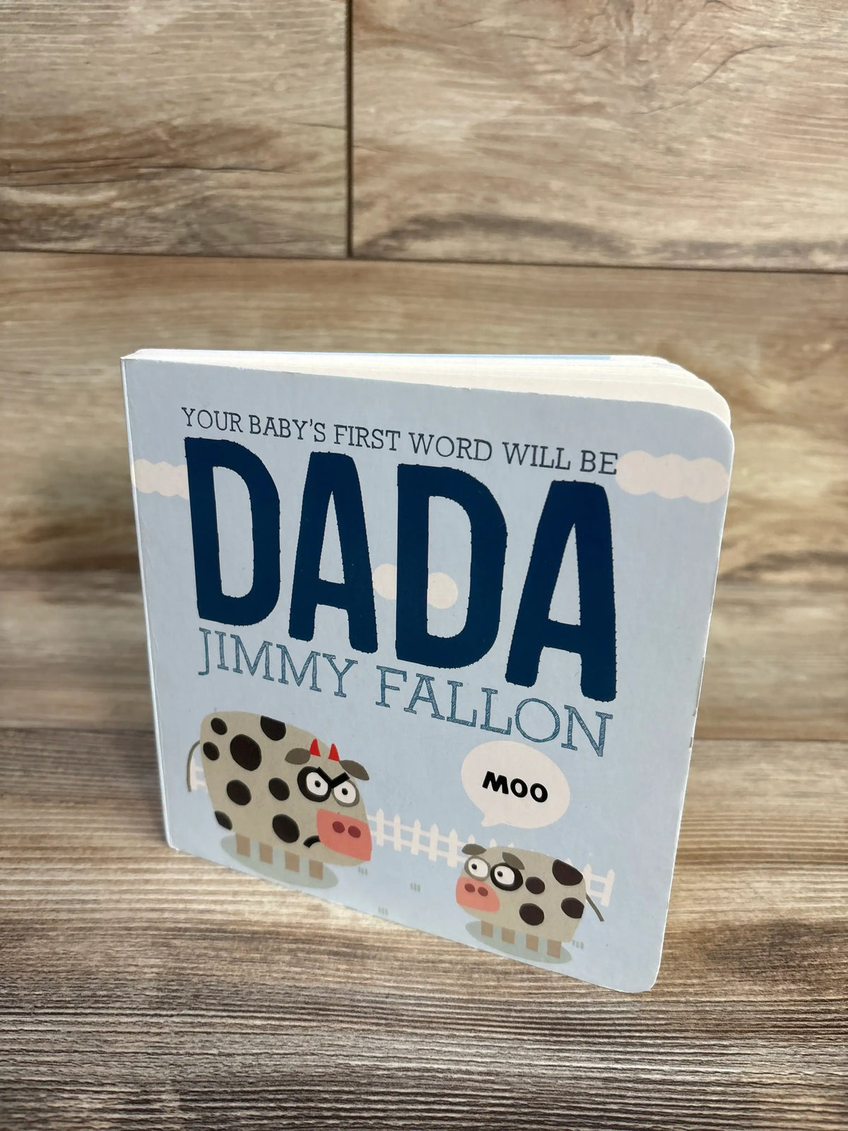 Front angled view of Your Baby’s First Word Will Be Dada Board Book By Jimmy Fallon, showing two cows and Moo speech bubble