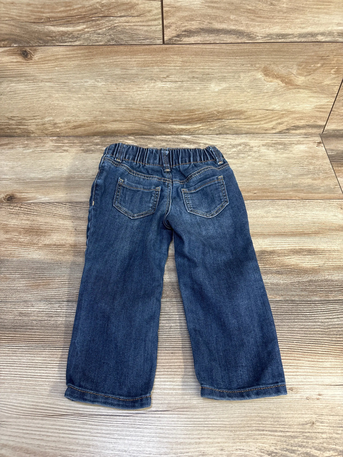 Rear view of OshKosh baby jeans in blue, size 18 months, showcasing elastic waistband and two back pockets on a wood-look floor