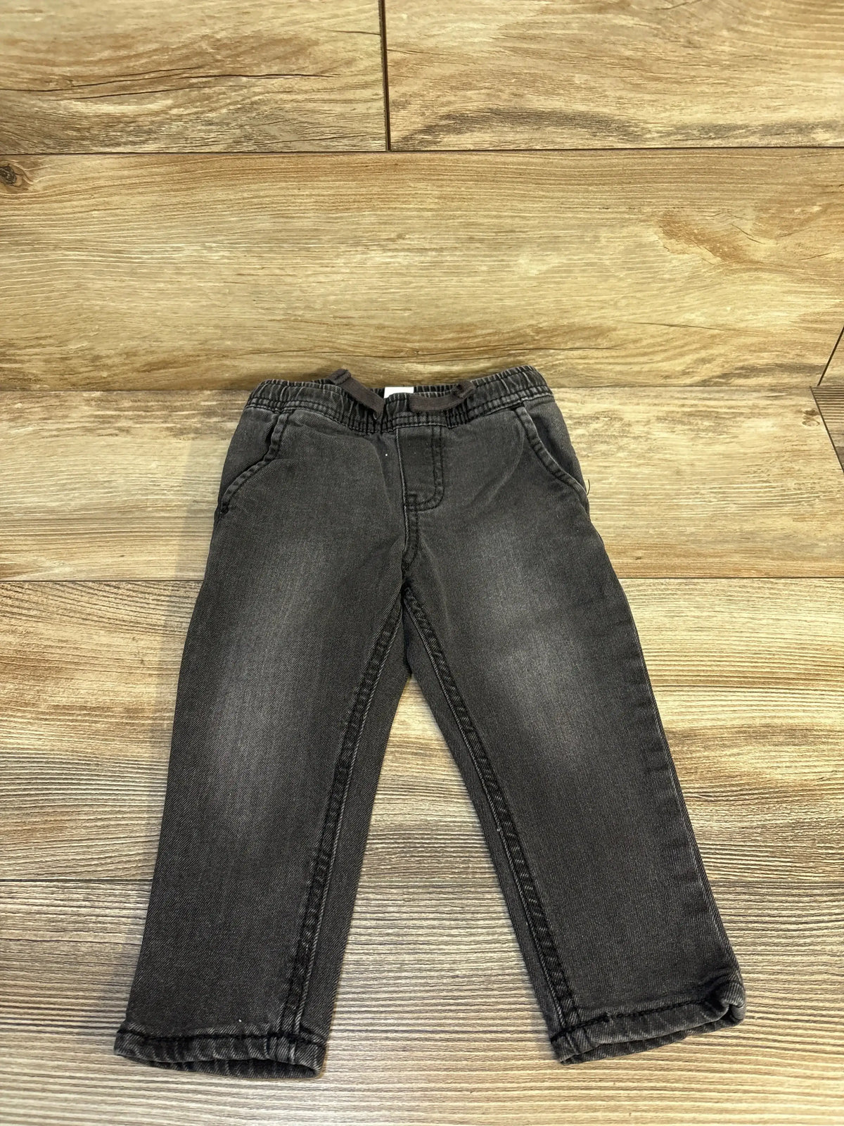 Flat lay of OshKosh toddler drawstring jeans, 18 months, showing elastic waistband and faded grey wash on wood floor