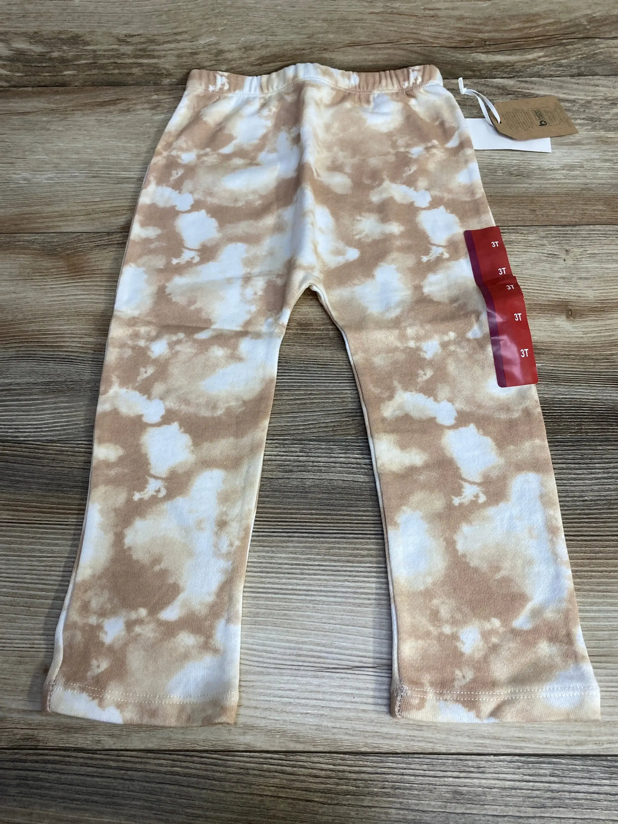 Top-down flat lay of toddler tie-dye pants (3T) showcasing beige and white cloud-like pattern, elastic waistband, and size tag on wood background