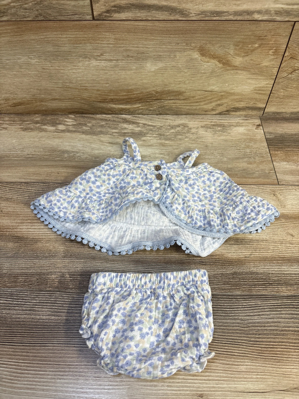 Flat lay of Rabbit Bear organic 2-piece blue floral tank top with wooden buttons and ruffle trim, and matching elastic-waist bloomers on wood surface