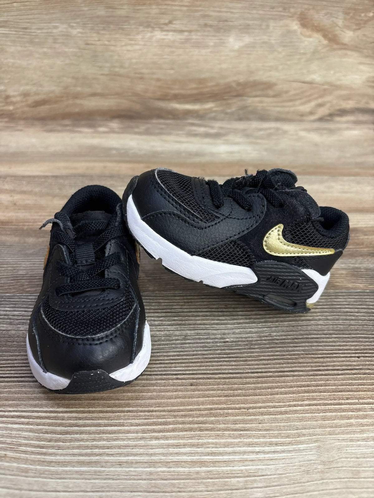 Black and white toddler Nike Air Max sneakers with gold swoosh on wooden floor